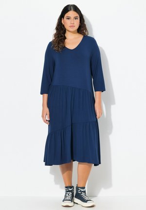 Marineblaues, knielanges Kleid mit V-Ausschnitt und dreiviertellangen Ärmeln. Es hat gestufte Rüschen am Saum und einen weichen, strukturierten Stoff.