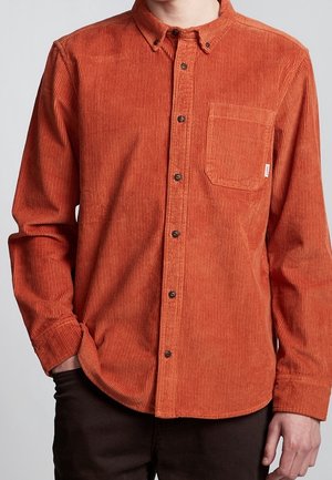 Mand iført en brændt orange kordbuks-skjorte med knapper og en brystlomme, stående med den ene hånd i lommen mod en ensfarvet baggrund.