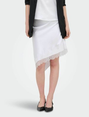 Femme portant une jupe asymétrique en satin blanc avec bordure en dentelle, un blazer noir et des talons pointus noirs, debout devant un fond uni.
