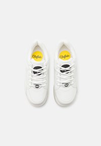 Bílé tenisky s hladkým koženým povrchem, kulatou špičkou, šněrováním a černo-žlutými logy na jazyku a stélce.