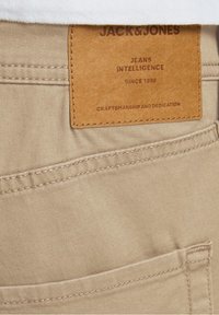 Los jeans de mezclilla beige cuentan con una etiqueta de cuero con texto en relieve: "Jack & Jones, Jeans Intelligence, Desde 1990, Artesanía y Dedicación."