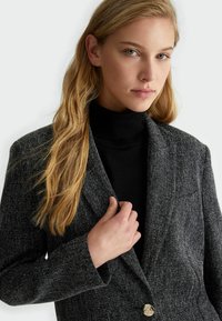Blazer a righe bianche e nere con un unico bottone dorato, rever a scollo, e vestibilità sartoriale, indossato sopra un maglione a collo alto nero.