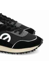 Chaussures en suede noir avec des accents gris, présentant un dessus texturé, un système de laçage et une semelle en caoutchouc avec des pointes pour une meilleure adhérence. Logo circulaire sur le côté.