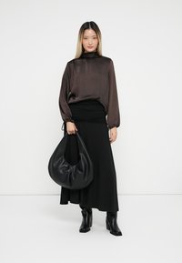 Long-sleeve, brown satin blouse with a high collar, paired with a black, fitted wrap skirt. Model holds a black, slouchy handbag. Black ankle boots.