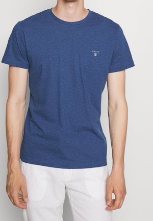 Man wearing a blue short-sleeve GANT t-shirt and white pants, standing against a plain light background.