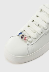 White leather sneaker with textured surface, featuring flat white laces and three jeweled lace accents in blue, red, and pink.