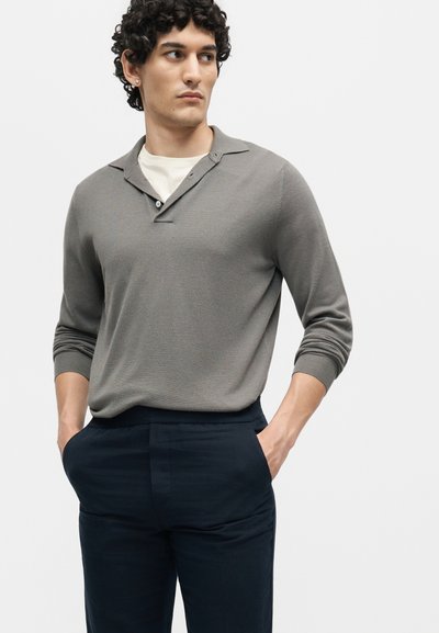 Νέος άνδρας με σγουρά μαλλιά που φορά γκρι polo με μακριά μανίκια πάνω από λευκό μπλουζάκι, σκούρο παντελόνι, με τα χέρια στις τσέπες, κοιτάζει αριστερά.