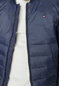 Námořnická puffy bunda se zipem vpředu a vroubkovaným límcem. Obsahuje prošívané panely a malé logo v červené, bílé a modré barvě na prsou.