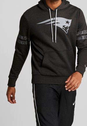 Osoba nosząca ciemnoszarą bluzę z kapturem New England Patriots z logo i tekstem na rękawach, w połączeniu z czarnymi spodniami Nike z białymi paskami.