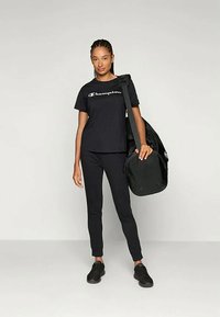 Žena oblečená v čiernom tričku Champion, čiernych nohaviciach a čiernych teniskách, nesúca veľkú čiernu športovú tašku cez plece.