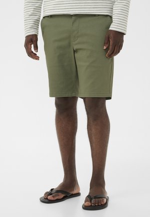 Mand iført olivengrønne knælange shorts, sorte klipklappere og en hvid langærmet skjorte med lysegrå striber.