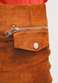 Närbild av en brun mockaskörtsficka med en horisontell silverdragkedja ovanför och en tryckknappsfåll nedanför.