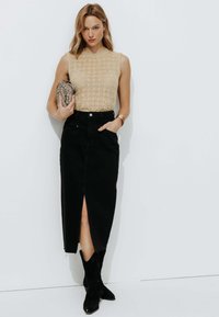 Haut sans manches beige en maille associé à une jupe maxi en denim noire avec une fente avant. La mannequin tient une pochette texturée et porte des bottines noires.