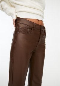 Detailní záběr na osobu, která má na sobě vysoké tmavě hnědé kožené kalhoty a krémový pletený svetr, ruka opřená o bok.