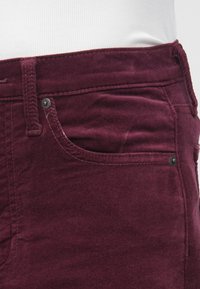 Pantaloni din catifea plisată mov închis, cu croială subțire, având cusături subtile, un buzunar frontal și închidere cu un singur nasture. Textură neagră vizibilă.