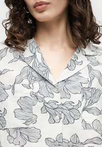 Γυναίκα με καστανά σγουρά μαλλιά μέχρι τους ώμους που φοράει λευκή μπλούζα με μαύρη floral γραμμική τέχνη και V-λαιμόκοψη.