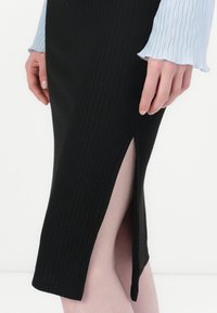 Jupe noire côtelée avec une fente sur le côté. Le tissu est texturé et ajusté, associé à une chemise à manches longues bleu clair avec des poignets bouffants.