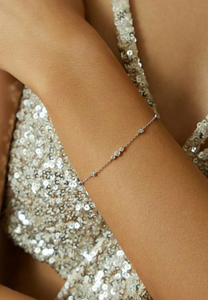 Silberne Gliederarmband mit kleinen runden Verzierungen mit strukturierten Oberflächen, getragen an einem gebräunten Arm vor einem glitzernden, paillettenbesetzten Hintergrund.