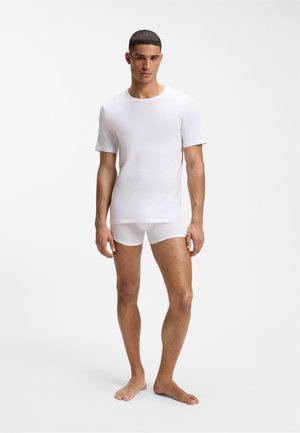 Man standing barefoot wearing a white short-sleeve t-shirt and white boxer briefs against a plain white background.