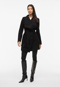 Abrigo negro de tela suave, con un cuello ancho, cintura ceñida con cinta y mangas largas, combinado con pantalones negros ajustados y botas hasta la rodilla.