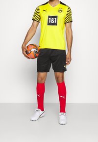 Gul och svart randig fotbollströja med logotyp, svarta shorts, röda strumpor och vita fotbollsskor; spelaren håller en texturerad orange fotboll.