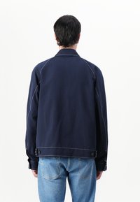 Mornarsko moder plašč iz teksturiranega blaga, ki ima belo šivanje, ovratnik in zapnete manšete, v kombinaciji s svetlo modrimi kavbojkami.