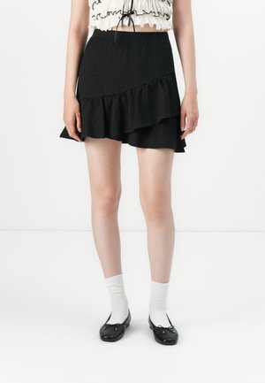 Kvinne iført et svart volangminiskjørt, hvite sokker, svarte ballettsko og en hvit cropped topp med svart kant, stående på et hvitt gulv.