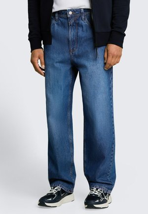 Uomo che indossa jeans blu larghi, sneakers nere e bianche, camicia bianca e giacca nera con zip su sfondo semplice.