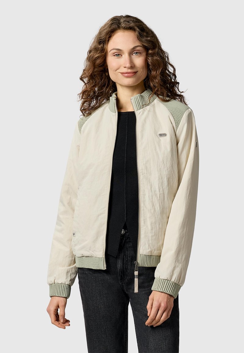 Jeune femme aux cheveux bouclés portant une veste zippée beige clair avec des poignets et un col côtelés, sur un haut noir et un jean foncé.