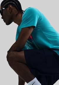 Camiseta de manga corta color azul verdoso con un diseño gráfico colorido en la parte frontal, combinada con shorts navy y calcetines blancos, usada por una figura sentada.