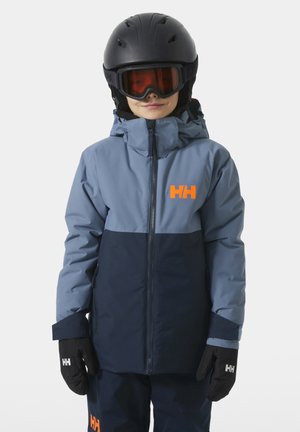 Kind trägt blaue und marineblaue Skijacke, schwarzen Helm, orangefarbene Skibrille und schwarze Handschuhe mit HH-Logos vor grauem Hintergrund.