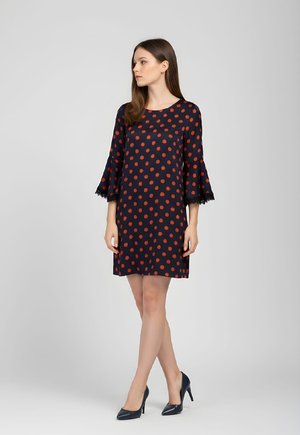 Mujer de pie con vestido azul marino con lunares rojos y mangas acampanadas con ribete de encaje, usando tacones negros, mirando hacia la izquierda.