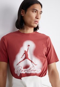 Piros pamut póló, fehér sziluettel egy ugró figuráról és „Jordan” felirattal írásos betűtípussal az alján. Rövid ujjú, kerek nyakkivágással.