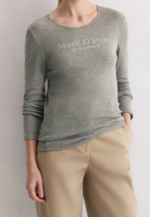 Femme portant une chemise à manches longues gris clair avec le texte « Marc O’Polo EST. IN STOCKHOLM » et un pantalon beige, debout, une main dans la poche.