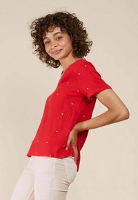 Haut rouge à manches courtes avec un col en V, arborant de subtils motifs triangulaires dorés. Fabriqué en tissu texturé, avec une coupe décontractée.