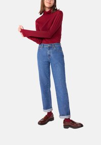 Maglione rosso a maniche lunghe con colletto alto, jeans in denim blu a vita alta con risvolti, mocassini marroni con una finitura lucida e calzini bordeaux.