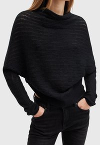 Femme portant un pull noir ample en côtes avec de longues manches ajustées et un jean noir, les mains partiellement dans les poches.