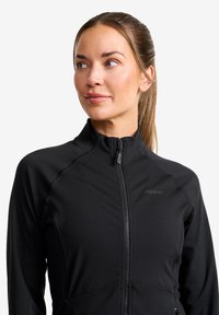 Svart träningsjacka med dragkedja och kroppsnära passform, som har en hög krage och platta sömmar. Tyget ser slätt och lättviktigt ut.