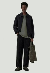 Black zip-up jacket, olive green t-shirt, black wide-leg pants, black shoes, carrying a quilted green bag with straps, standing against a light background.