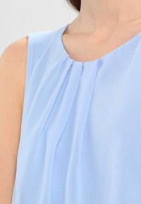 Blusa sin mangas de color azul claro con cuello redondo, que presenta suaves pliegues y una textura de tela suave y fluida.