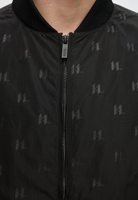 Gros plan d'une veste noire avec une fermeture éclair et un motif subtil répétitif "KL", montrant le cou et la partie supérieure de la poitrine.
