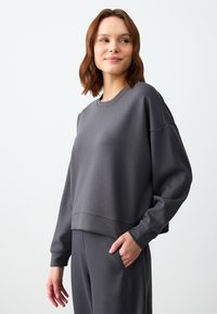 Felpa grigia corta con scollo rotondo, spalle abbassate e polsini a costine, abbinata a pantaloni coordinati. Tessuto in morbido misto cotone.