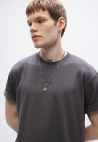 Camiseta de algodón gris con las mangas remangadas, que presenta un logo "BOSS" en relieve. Se lleva un collar de cadena plateada con un colgante de brújula.