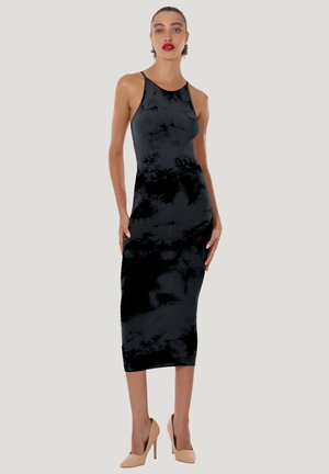 Kvinna i ärmlös svart tie-dye midiklänning och nudefärgade högklackade skor, stående med händerna vilande på höfterna mot en enfärgad bakgrund.