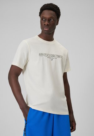 Mężczyzna o ciemnej skórze, ubrany w białą koszulkę "SPORTSWEAR" i jasnoniebieskie spodenki, stojący z jedną ręką w kieszeni, patrzący na bok.