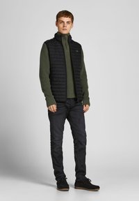 Sort puffer vest over en mørkegrøn striktrøje, parret med mørke jeans og sorte ruskinds sko. Simpelt design med en lynlås kravedetail.