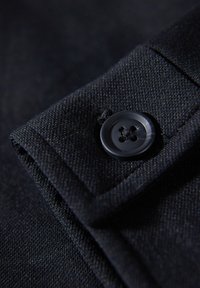 Gros plan d'un poignet en tissu noir sécurisé par un bouton noir mat à quatre trous, montrant un matériau tissé texturé et des détails de couture.
