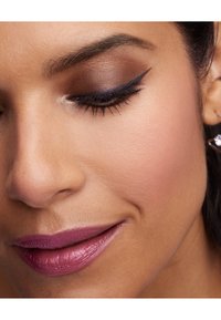 Nærbilde av et ansikt med glatt, solbrun hud, myk brun øyenskygge, kraftig eyeliner og en dempet mauve leppestift med litt glans.