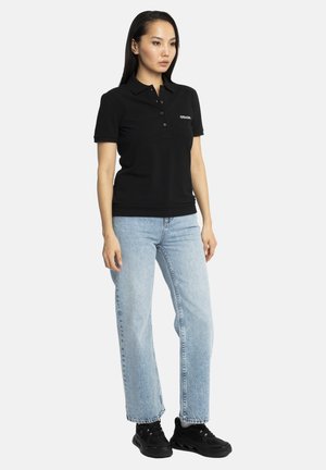Femme aux longs cheveux foncés, portant un polo noir, un jean bleu clair et des baskets noires, debout devant un fond uni.