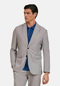 Blazer grigio chiaro su misura con rever a punta, due tasche frontali e bottoni neri, indossato sopra un polo blu con rifiniture bianche.